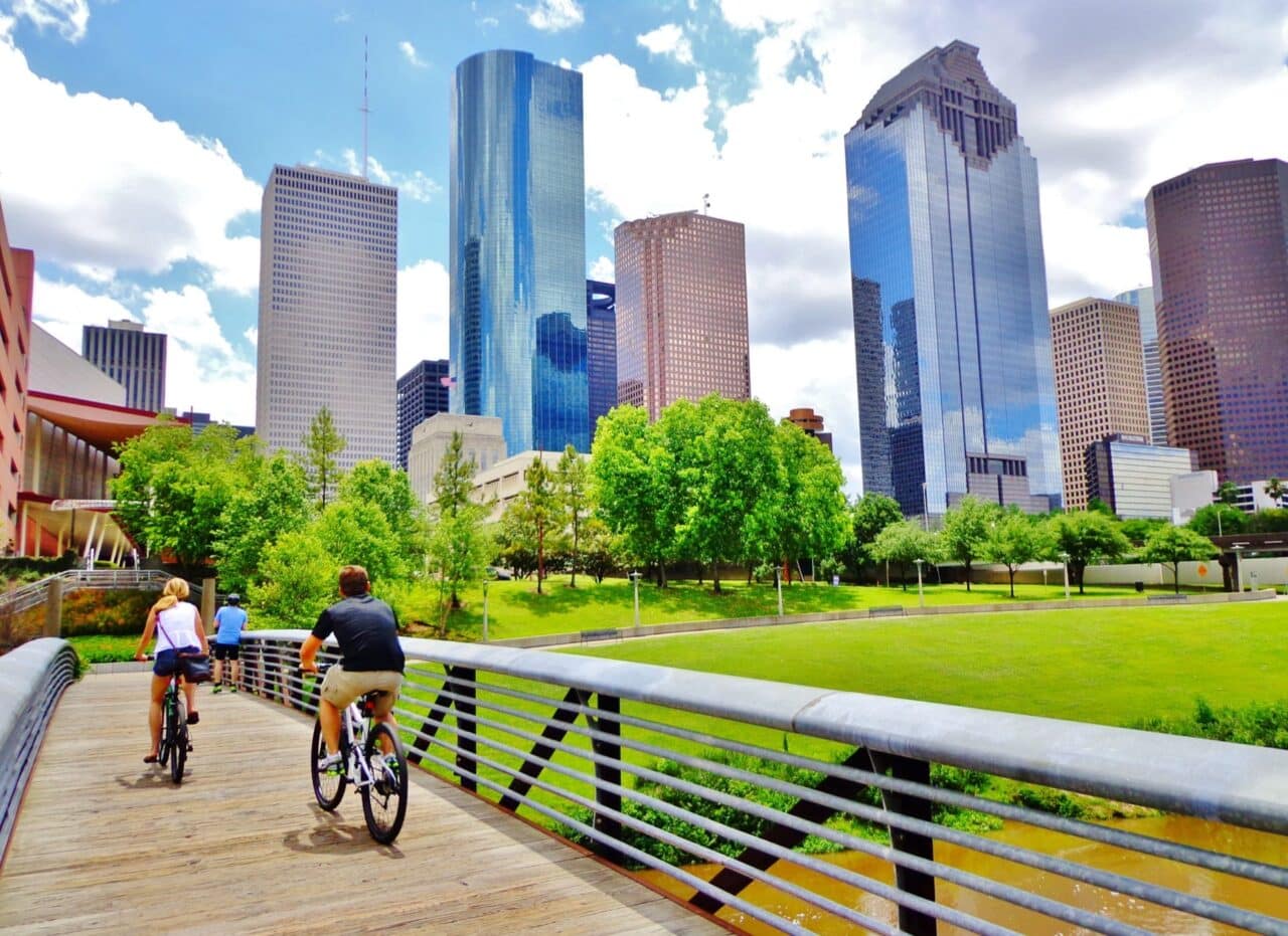 Bicyclists cross wooden bridge in buffalo bayou park, with a beautiful view of downtown houston (skyline / skyscrapers) in background on a summer day - houston, texas, usa